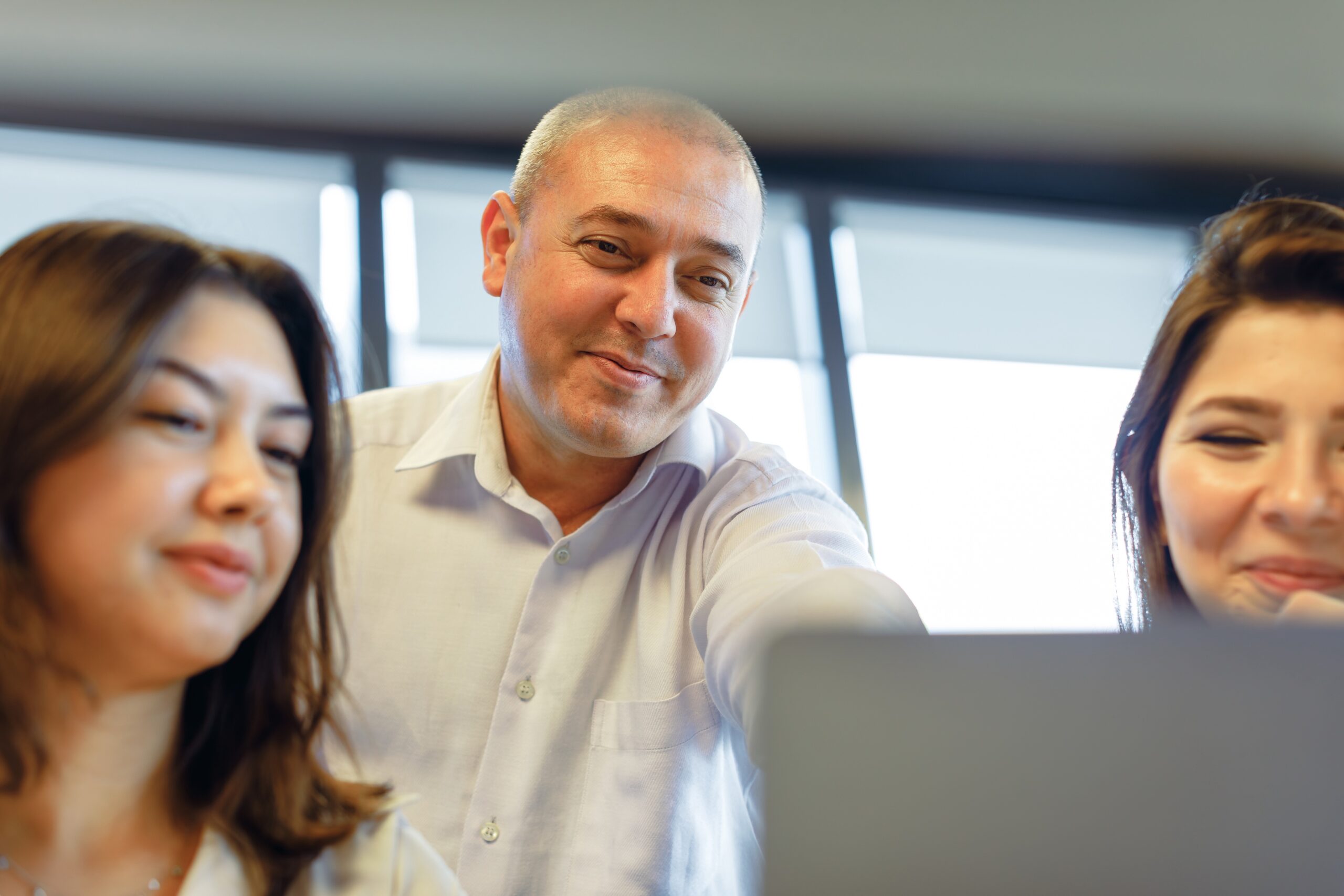 Portrait of happy creative business teamwork on a desktop computer and looking monitor screen. The manager businessman point on the screen to young female business workers while standing in a modern office meeting room Joyful Collaboration: Manager Guides Young Professionals in Modern Office. Immerse yourself in the vibrant energy of a modern office as a team of happy business people gathers around a computer, their faces beaming with enthusiasm. The middle-aged manager, a seasoned boss, takes center stage, passionately showing and explaining valuable insights on the screen to two young, beautiful businesswomen or interns. Witness the exchange of information and knowledge flow seamlessly among working business employees, creating an atmosphere of shared success and growth in the workplace